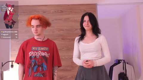 Video of two people: a man with red hair in a Spider-Man shirt and a woman with long black hair in a white top. Background includes wooden wall and purple lighting.
