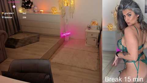 Video of a curvaceous woman with long, dark hair, wearing a green, sheer lingerie set, posing seductively in a cozy, dimly lit room.