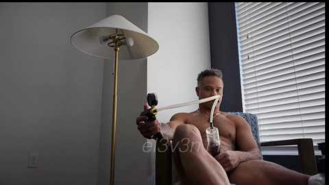 Video of a nude, muscular, middle-aged man smoking a hookah pipe, sitting in a chair beside a white lamp and window with closed blinds in a minimalist room.