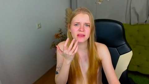 Video of a slender, fair-skinned, topless, blonde woman with long hair, wearing a distressed expression, with a small tattoo on her left forearm, sitting in a black and beige chair in a room with a green couch and light gray walls.