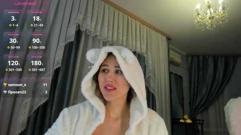 A video of a light-skinned woman with a medium build, wearing a fluffy white robe with a bear hood, standing indoors in a dimly lit room with gray curtains and a chandelier.