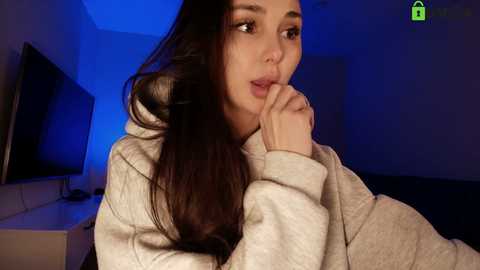 Video of a young woman with long brown hair, wearing a gray hoodie, sitting pensively, blue lighting, simple bedroom background, dim lighting, close-up.