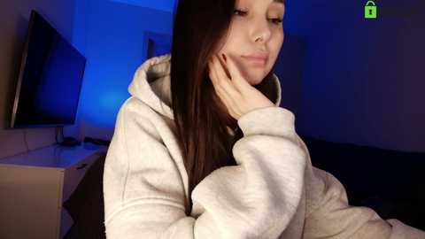 A video of a young woman with straight, dark hair, wearing a light-colored hoodie, resting her chin on her hand in a dimly-lit room with a blue LED light.