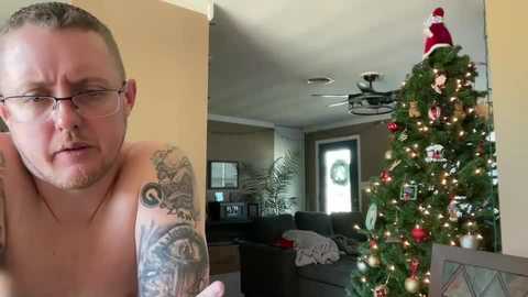 Video of a shirtless man with glasses and tattoos, gazing at a decorated Christmas tree in a cozy living room with beige walls.