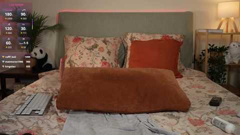 Media: A video of a cozy bedroom with a floral-patterned bedspread, two pillows, a beige blanket, and a wooden nightstand with a lamp and a teddy bear. A digital clock and a remote control are on the bed.