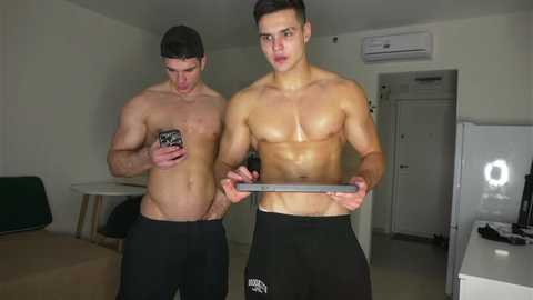 Video of two shirtless, muscular men with light skin and short hair, both in black pants, holding smartphones. Background includes white walls, air conditioning unit, and a TV.