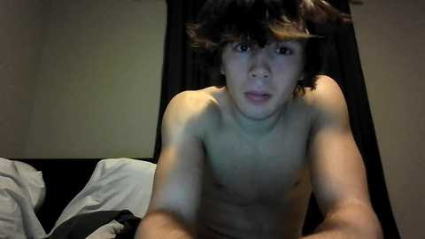 Video of a young, shirtless, slim Asian boy with tousled black hair and a slightly open-mouthed expression. He is lying on a bed with white sheets, against a dark curtain background.