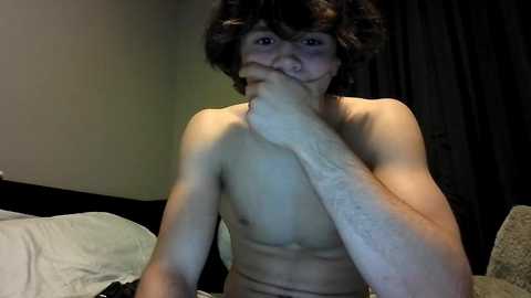Video of a young, shirtless man with short, curly hair, covering his mouth with his right hand, sitting on a bed with white sheets, in a dimly lit room with dark curtains.