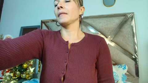 Video of a woman in a maroon ribbed sweater, slightly blurry, standing in a room with a Christmas tree and a large, shiny, silver headboard.