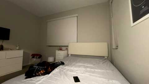 Video of a minimalist, dimly-lit bedroom with a white bed, beige walls, a window with a white blind, a white dresser, and a black TV on the dresser.