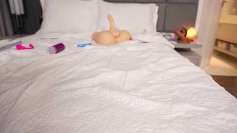Video of a neatly made bed with white sheets, featuring a realistic, lifelike, uncircumcised penis-shaped pillow placed centrally. Pink and white vibrators lie nearby, and a bedside lamp casts a warm glow.