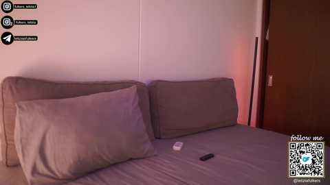Video of a dimly lit bedroom with a beige bed and two large pillows, a TV remote on the bed, and a QR code in the bottom right corner.