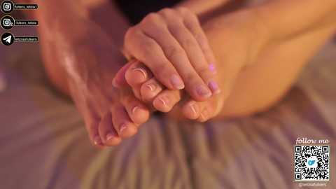A video of a close-up of a woman's feet, with neatly trimmed nails, being held and massaged by another pair of hands. The background is blurred, suggesting a cozy setting.