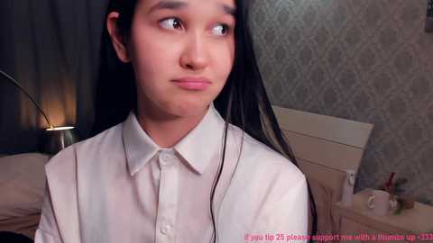 Video of a young woman with long black hair and fair skin, wearing a white blouse, looking contemplative in a bedroom with a patterned wallpaper and a nightstand with a lamp.