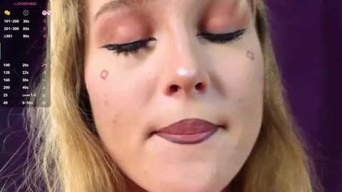 Video of a young woman with fair skin, closed eyes, and subtle makeup, including a small mole near her right eye. Background is dark purple.