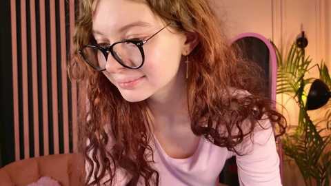 Video of a young woman with curly brown hair, wearing black-rimmed glasses and a light pink top, smiling in a cozy room with a pink tufted couch, green plant, and modern decor.