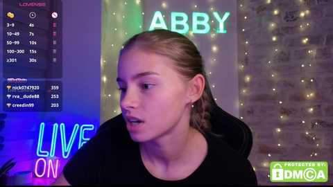 Video of a fair-skinned, young woman with long blonde hair in braids, wearing a black top, speaking into a camera in a dimly lit room with string lights and \"LIVE ON\" neon sign.