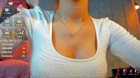 Video of a woman in a white, long-sleeve, ribbed-knit top with a low neckline, showcasing her cleavage. She wears a dainty necklace. Background shows a cozy, warm-toned room with a plush, pink couch.