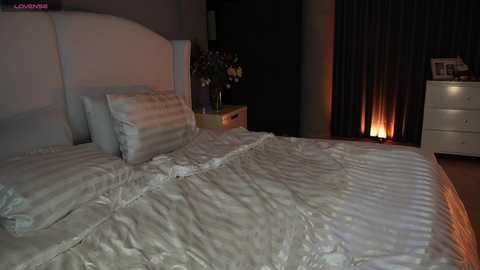 Video of a dimly lit bedroom with a white bed, striped pillows, and a white headboard. Nightstand with flowers and framed photos. Dark curtains and a lit candle create a cozy atmosphere.