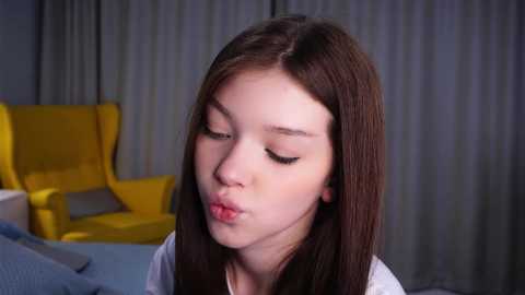 Media: A video of an Asian woman with long, straight brown hair, wearing a white top, making a pouty kiss in front of a gray curtain background with a yellow armchair and blue bed.