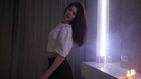 Video of a young Asian woman with long black hair, wearing a white crop top and black skirt, standing in a dimly lit room with candles on a white counter, against a grey curtain.