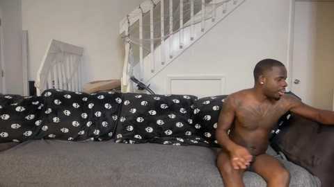 Video of a shirtless Black man with a muscular build, sitting on a gray sofa with black cushions featuring white skull patterns. The background shows a white staircase and white door.