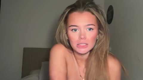 Video of a young Caucasian woman with long, straight blonde hair, fair skin, and light blue eyes. She is topless, with a neutral expression, in a dimly lit, plain room.