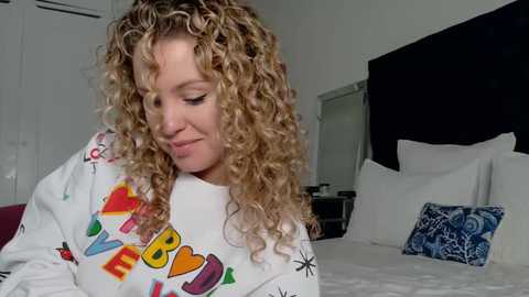 Video of a curly-haired woman in a colorful \"LOVE\" sweatshirt, smiling while looking down, in a modern, white bedroom with a black headboard and blue patterned pillow.