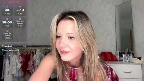 Video of a blonde woman in a pink dress, smiling in a bedroom with a mirror, clothes, and a digital screen displaying social media metrics.