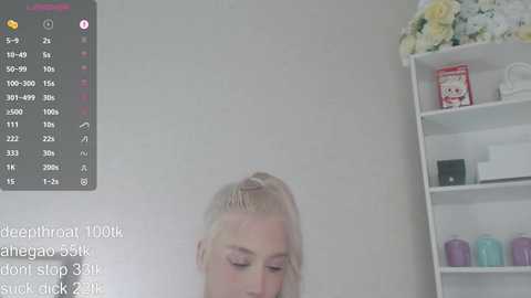 Video of a blonde woman with light skin, wearing makeup, standing in a minimalist, white-walled room. She has her hair styled in a high ponytail. A digital overlay shows a chat with the text, \"Hello, I'm Daniella, don't slip 33.5 suck check.\