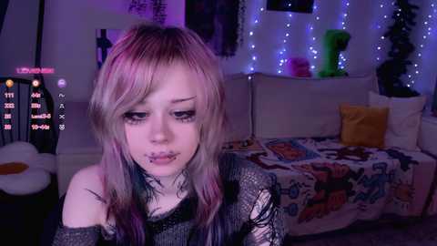 Video of a pale, slender young woman with long, multicolored hair, wearing black lace, in a dimly lit, whimsical room with fairy lights and colorful pillows.
