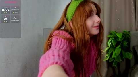 Video of a young woman with long red hair and a green headband, wearing a pink sweater, smiling while taking a selfie in a modern, light-filled room with a green plant.