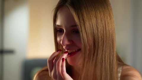 Video of a young, fair-skinned woman with long, straight blonde hair, wearing a white tank top, smiling while eating red food. Soft, warm lighting in a blurred, beige-toned background.