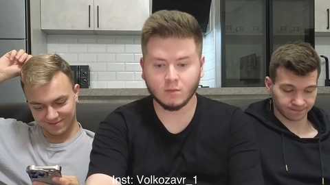 Video of three young men with short hair, wearing casual black t-shirts, sitting in a modern kitchen. The man in the middle has a large, square-shaped head.