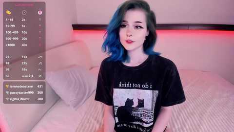 Media: Video of a young woman with blue hair, wearing a black t-shirt featuring a cat graphic, sitting on a bed in a dimly lit room.