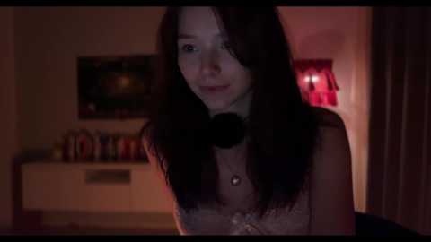 Video of a young woman with straight, shoulder-length dark hair, wearing a black choker necklace, standing indoors. Background features a dimly lit room with a TV and bookshelves.