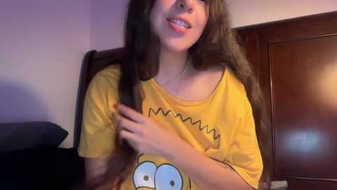 Video of a young woman with long brown hair, fair skin, and wearing a yellow SpongeBob T-shirt, smiling and playfully pulling her hair in a bedroom with a wooden door and purple wall.