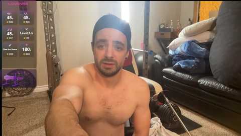 Video of a shirtless, muscular man with a dark beard, wearing a backward cap, sitting in a cluttered living room with a video game screen in the background.