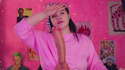 Media: Video of a young woman with pale skin, dark hair, and large breasts, wearing a pink shirt, holding her head, with a large erect penis in front of her. Background features a pink wall adorned with posters.