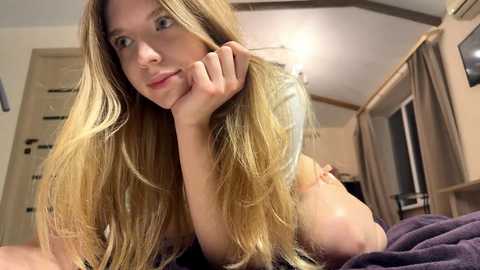 A video of a young Caucasian woman with long blonde hair, light skin, and blue eyes, lying on her stomach, resting her chin on her hand, in a softly lit bedroom.
