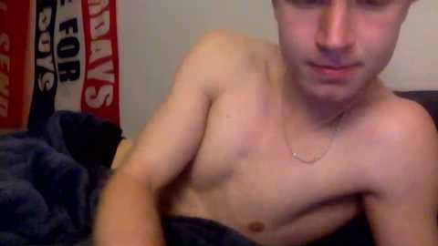 Video of a shirtless young man with fair skin and light brown hair, wearing a silver chain necklace, leaning on a dark blanket, in a dimly lit room with a red and white banner in the background.