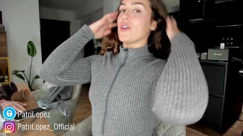 Video of a woman with medium brown skin, wearing a gray knit sweater, adjusting her hair. Background features a modern living room with dark furniture and a green plant.