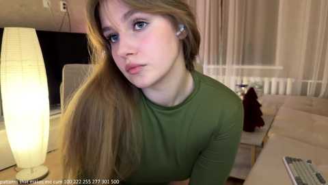 Media: Video of a young Caucasian woman with long, straight blonde hair, wearing a green top, looking pensive in a modern living room with a beige couch, white radiator, and a lit floor lamp.