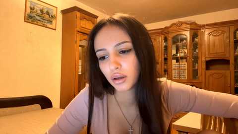 Video of a young woman with long dark hair, wearing a light pink cardigan, in a warm-toned, wooden kitchen with cabinets and framed landscape art on the wall.