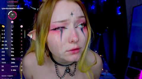 Media: Video of a young, pale-skinned woman with blonde hair and pointed ears, wearing a black choker and star tattoos on her face, in a dark, dimly lit room.