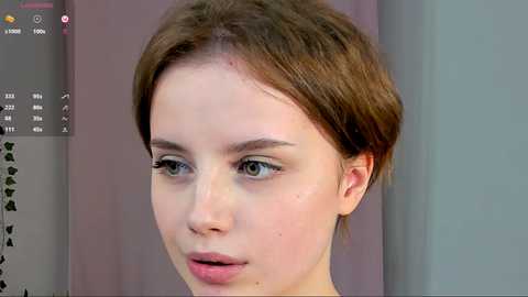 Media: A close-up video of a young woman with short brown hair, fair skin, and blue eyes, wearing minimal makeup. She has a neutral expression and is indoors against a plain background.