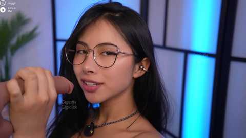 Video of an Asian woman with long black hair and glasses, wearing a necklace, holding a large, erect penis, against a blue-lit backdrop.
