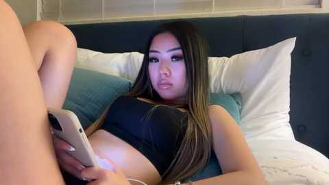 Media: Video of an East Asian woman with long, straight hair, wearing a black crop top, lying on a bed with white sheets, holding a smartphone, in a modern bedroom with white blinds and a dark headboard.
