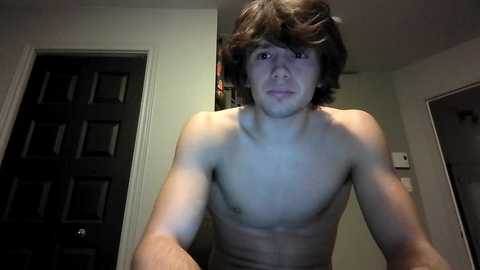 Video of a shirtless, young, slender Caucasian male with messy brown hair, light skin, and a neutral expression, standing in a dimly lit hallway.