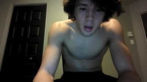 Video of a young, shirtless, fair-skinned man with messy, dark hair, leaning forward, mouth open, indoors against a beige wall with a dark door and light switch.